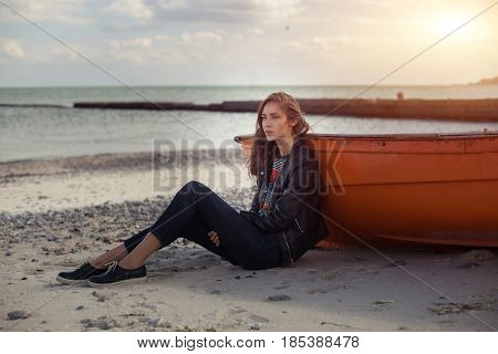 A girl sideways sitting near a red boat on the beach by the sea with beautiful sunset on the background. Woman in jeans pullover and jacket on the sand