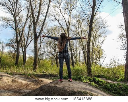 A young girl with long blond hair dressed in navy blue pants and pink sneakers stands on the hillock face to the sky. She spread her arms and enjoys the fresh air in the forest and enjoys life