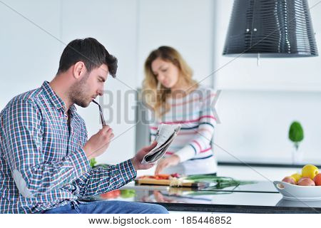 Couple standing and sitting at the kitchen while smiling and man reading a newspaper and holding mug before work