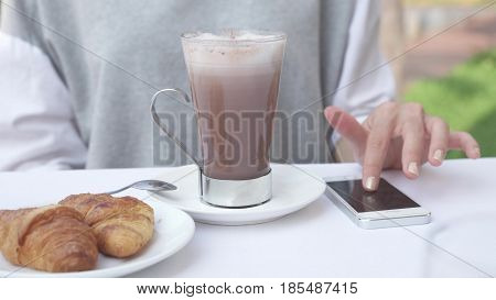 Young woman checks notifications on smartphone while drinking cacao with croissant. Female use mobile phone for business working during morning breakfast with hot chocolate. 4K