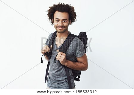 Confident and happy african young man with backpack smiling looking at camera ready to go hitchhiking or just hiking in mountains. White background. Copy space.