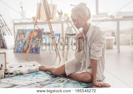 Beautiful young woman sitting on the floor in a bright Studio and paints with paint on canvas.
