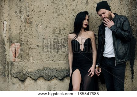 Pretty fashion woman or girl with long brunette hair in sexy black dress and bearded man in stylish jacket and hat smoking cigarette with wine bottle on grungy cement wall. Bad habits copy space