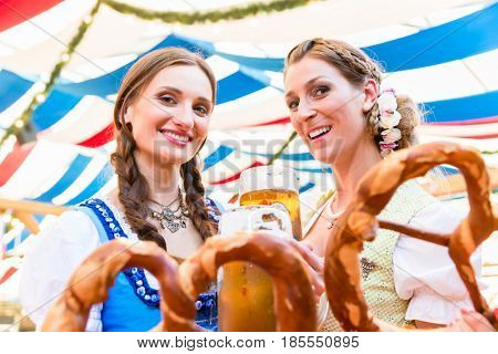 Two friends wearing dirndl are standing in a beer tent at Dult or Oktoberfest with giant pretzels