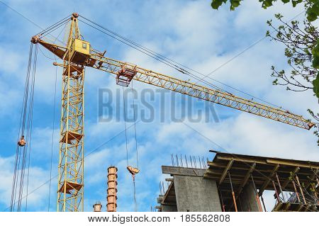 Yellow construction cranes and unfinished building with metal-concrete structure on a background of blue sky with white clouds. In the upper right corner of the green branches of a tree. Horizontal photo.