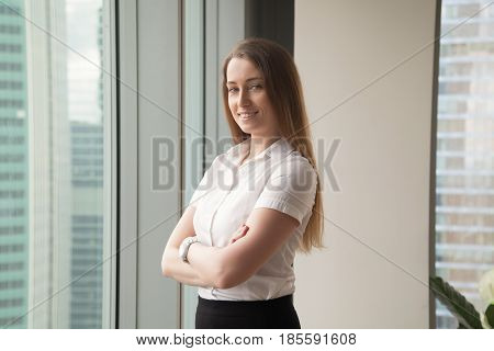 Confident businesswoman enjoys success. Happy woman standing in office with arms crossed, looking at camera. Smiling ambitious female entrepreneur thinking about business, feeling positive. Portrait