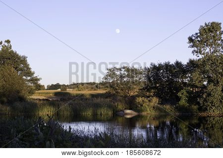 Landscape scene of a small river shoreline with trees foliage and reflections in the water near Windsor, Ontario with the moon visible on a sunny late afternoon in September.