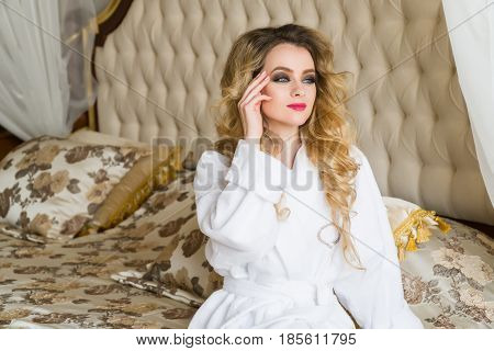 Beautiful seductive woman flirting with the camera sitting on a bed in White bathrobe looking up with a coquettish look.