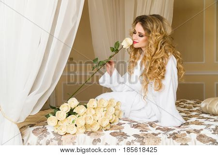 Beautiful seductive woman flirting with the camera is sitting on the bed with a big bouquet of white roses in White bathrobe looking up with a coquettish look.