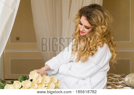 Beautiful seductive woman flirting with the camera is sitting on the bed with a big bouquet of white roses in White bathrobe looking up with a coquettish look.