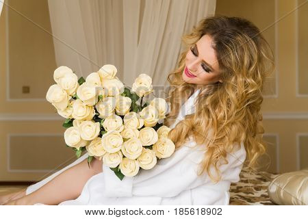Beautiful seductive woman flirting with the camera is sitting on the bed with a big bouquet of white roses in White bathrobe looking up with a coquettish look.