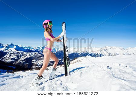 Side view portrait of female skier in a good shape standing in pink bikini and helmet on mountain peak