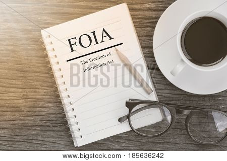 Concept FOIA (The Freedom of Information Act) message on notebook with glasses pencil and coffee cup on wooden table.