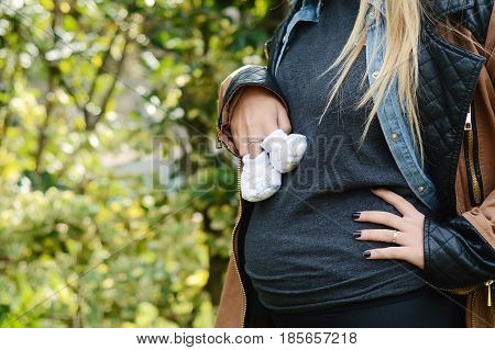 Portrait of beautiful young pregnant woman holding baby booties on her abdomen. Outdoors.