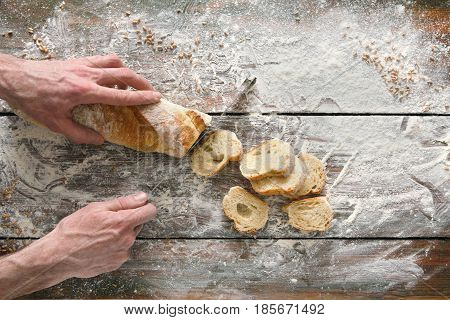 Baking background. Hands of baker cutting bread loaf with knife on rustic wooden table sprinkled with flour.