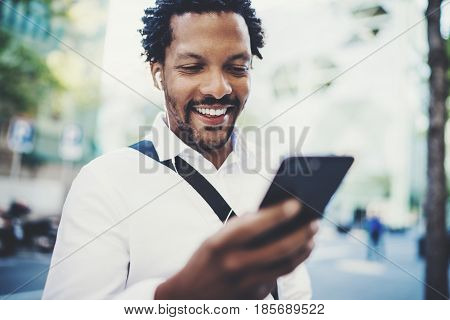 Closeup view of Happy american African man using smartphone outdoor.Portrait of young black cheerful man texting a sms message while listining to music.Blurred background, horizontal