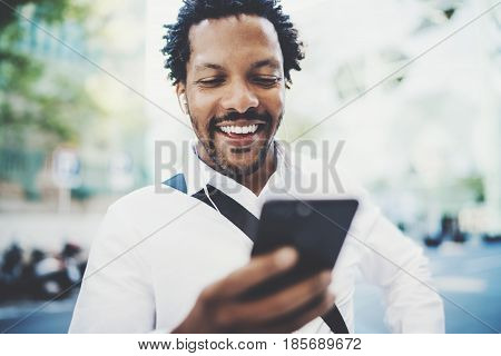 Closeup view of Happy american African man using smartphone outdoor to texting a sms message with friends.Portrait of young black cheerful man listining to music.Blurred background, horizontal