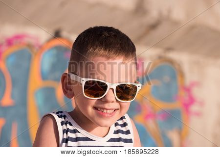 smiling small kid wearing sunglasses with sea sunset reflection in it and sailor shirt on graffiti background in summertime