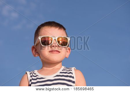 smiling little boy wearing white sunglasses with sea sunset reflection and sailor stripes vest on blue sky background with copyspace