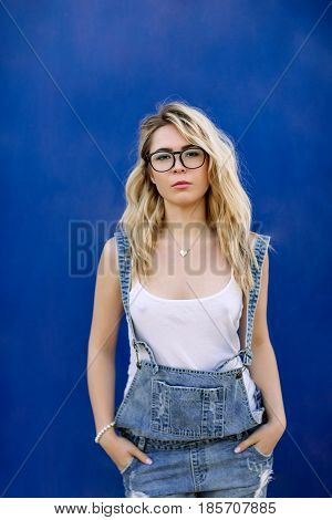 Young Stylish Blond Girl In Denim Overalls Made Of Denim And On A Blue Background