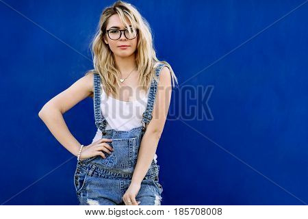 Young Stylish Blond Girl In Denim Overalls Made Of Denim And On A Blue Background