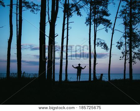 Silhouette young healthy man standing in wild forest rising hands up with beautiful sea sunset background
