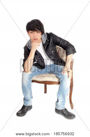 A young Asian man in a black leather jacket sitting in an old armchair bending forwards with hand on chin isolated for white background.