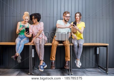 Multi ethnic group of young people dressed in colorful t-shirts chatting with gadgets sitting in a row indoors on the wall background