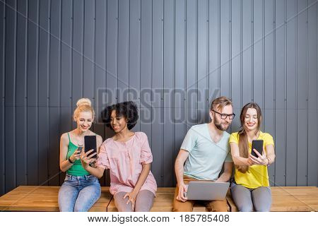 Multi ethnic group of young people dressed in colorful t-shirts chatting with gadgets sitting in a row indoors on the wall background