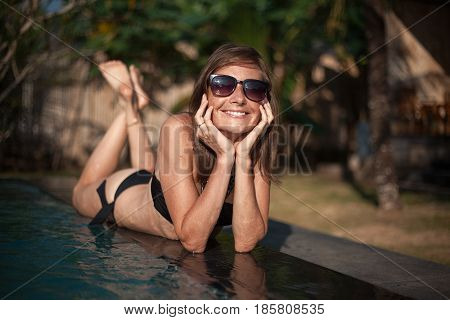 Horizontal portrait of smiling beautiful girl in black bikini and sunglasses lying on the pool edge