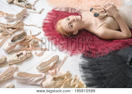 Attractive ballerina lies on the colorful tutus on the white floor in the studio. She wears a light top and holds pointe shoes. On the left there are many ballet shoes. Closeup. Horizontal.