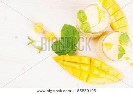 Freshly blended yellow mango fruit smoothie in glass jars with straw mint leaves mango slices top view. White wooden board background copy space.