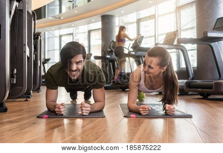 Cheerful young couple exercising forearm plank side by side on mats during workout in the interior of a modern fitness club