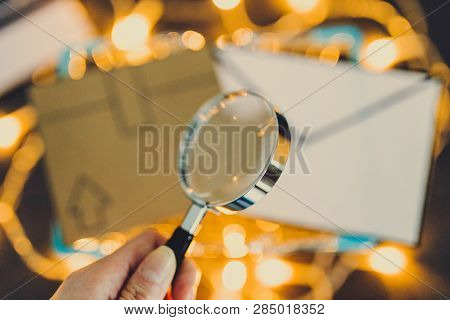 Hand Holding Magnifying Glass Over Shopping Cart With Cardboard Parcel And Email Envelope On Concret