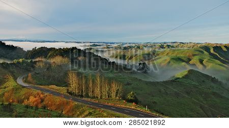 Road Leading Through The Green Hills With Clouds Rolling Over Them During The Sunrise On North Islan