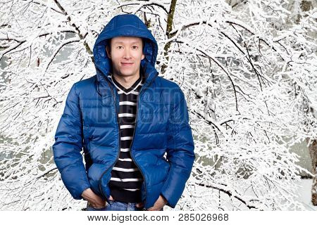 Happy Asian man in blue down padded coat, against snowy winter background. Man outdoors in warm winter clothes. Snow and cold