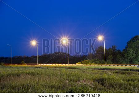 Lights Of Lanterns Near Highway. Night Scene. Grass In A Field At Front Plan. Trees In Woodland Belt