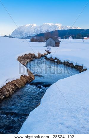 Small Curved Creek River With Bushes On Bank Winds Through A Snowy Field With Mountain Grimming In S
