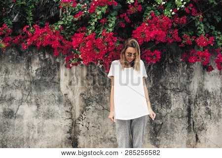 Hipster girl wearing blank white t-shirt posing against gray street wall with spring flowers, blank mockup for tshirt print store