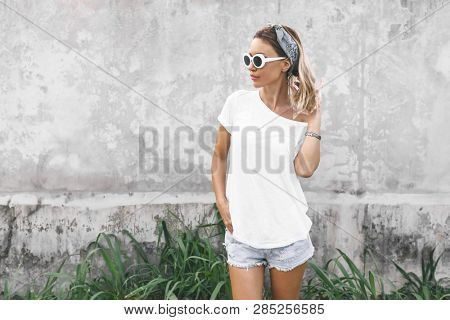 Hipster girl wearing blank white t-shirt and denim shorts posing against gray street wall, blank mockup for tshirt print store
