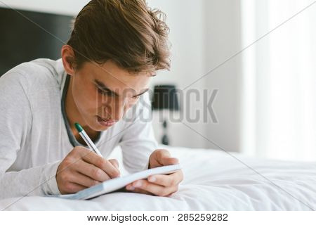 16 years old teenage boy writing school notes or doing homework while relaxing on couch in his room