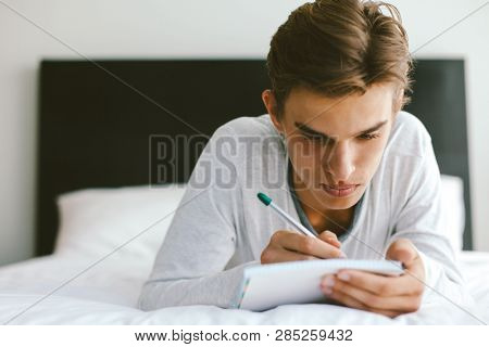 16 years old teenage boy writing school notes or doing homework while relaxing on couch in his room