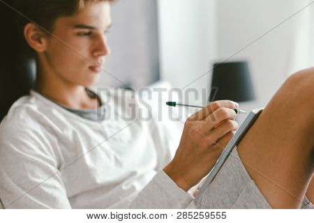 16 years old teenage boy writing school notes or doing homework while relaxing on couch in his room