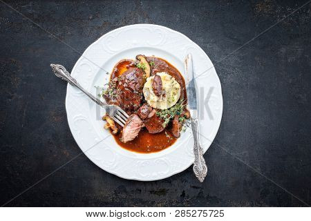 Traditional German braised pork cheeks in brown red wine sauce with mushroom and mashed potatoes as closeup on a classic design white plate 