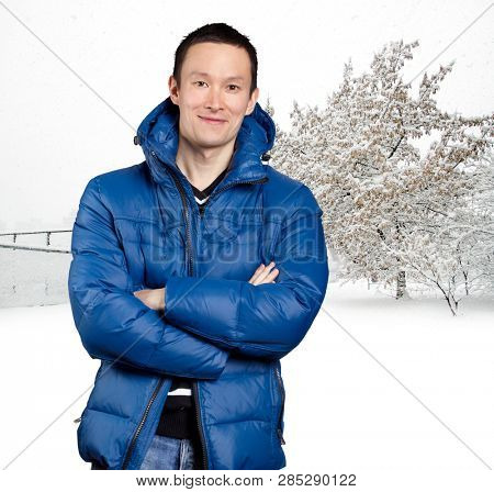 Happy Asian man in blue down padded coat, against snowy winter background. Man outdoors in warm winter clothes. Snow and cold