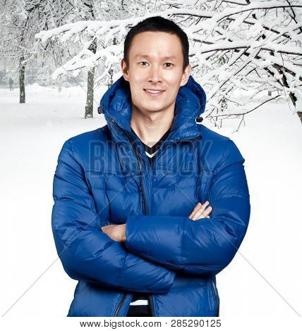 Happy Asian man in blue down padded coat, against snowy winter background. Man outdoors in warm winter clothes. Snow and cold