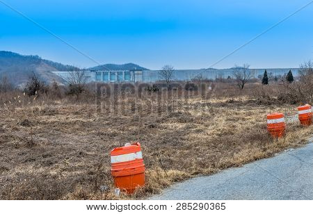 Orange And White Traffic Barrels In Field Of Dry Grass And Trees At Edge Of Rural Road With Large Co