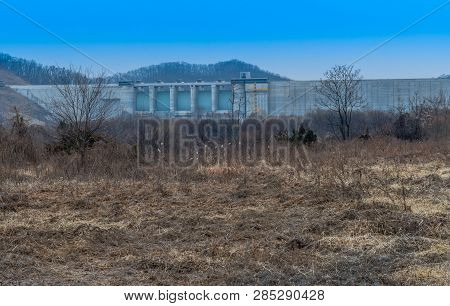 Winter Landscape Of Field Of Dry Grass And Trees In Front Of Concrete Dam Under Blue Sky.