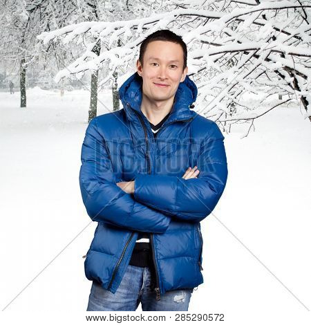 Happy Asian man in blue down padded coat, against snowy winter background. Man outdoors in warm winter clothes. Snow and cold