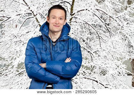 Happy Asian man in blue down padded coat, against snowy winter background. Man outdoors in warm winter clothes. Snow and cold